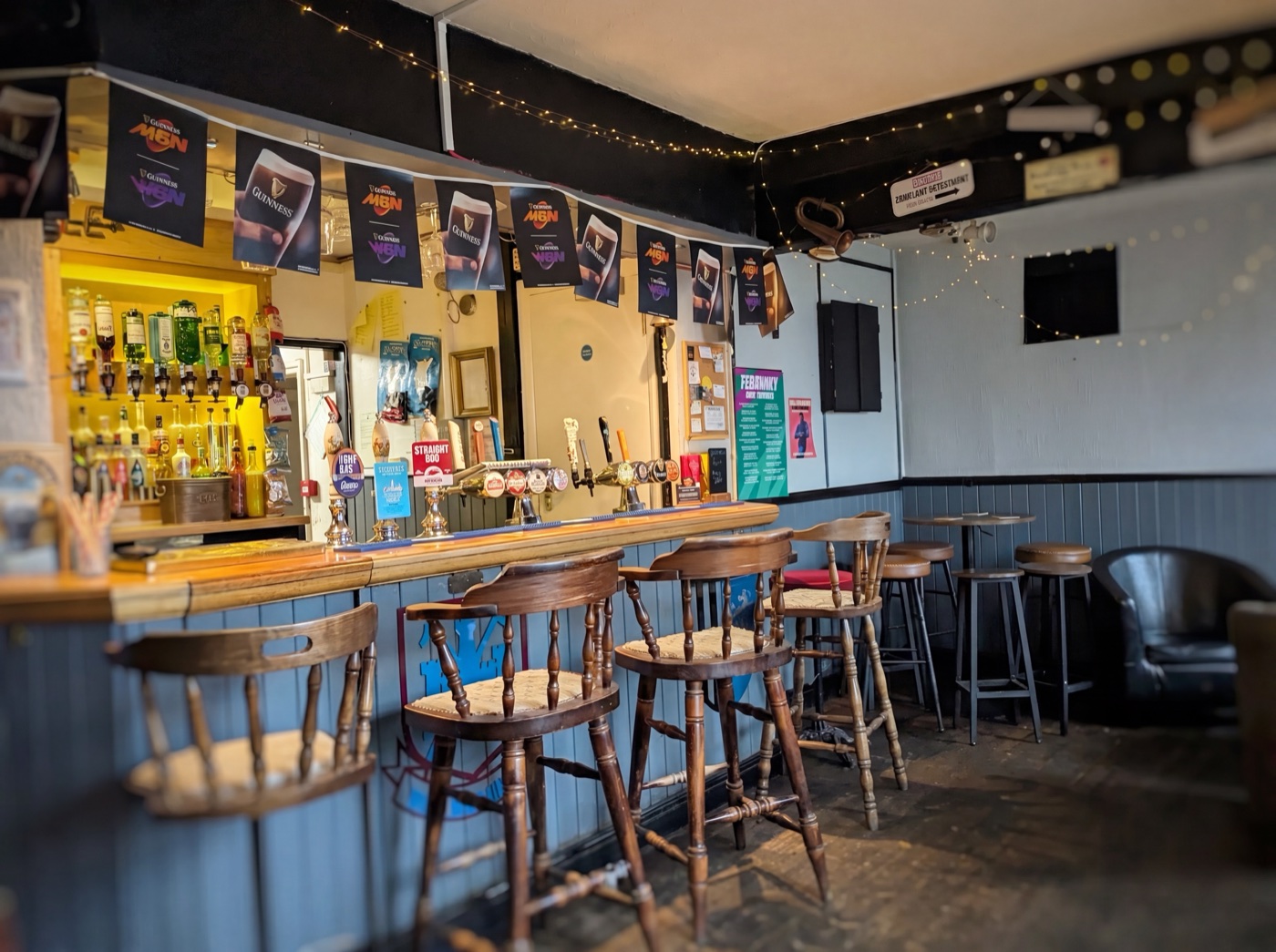 The bar at The Maypole Inn — wooden bar counter and stools, real ale pumps and spirit optics, with fairy lights and a leather tub chair